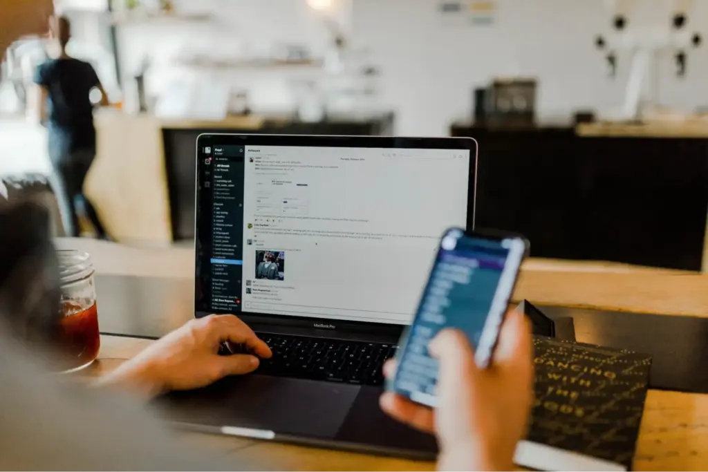 A person holding their phone in one hand while communicating with coworkers on their laptop messaging platform. 