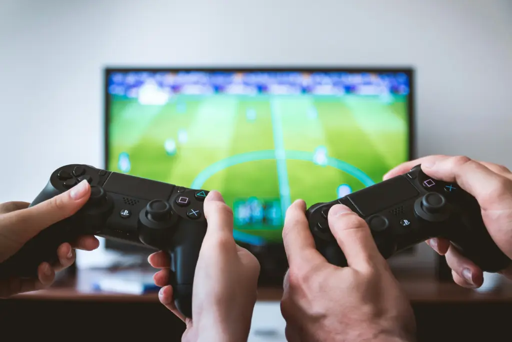 Two people holding playstation controllers with a tv in the background.